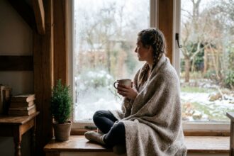 Femme en pull en laine assise sur un rebord de fenêtre avec une tasse de thé, contemplant un jardin givré en hiver, pause visuelle et bien-être hivernal